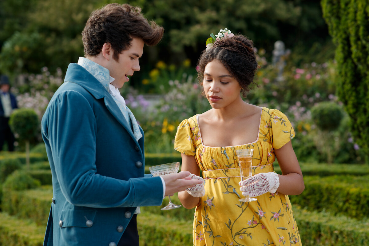 Two people in Regency-era clothing holding drinks, standing in a formal garden with greenery and flowers, engaging in conversation at an outdoor event.