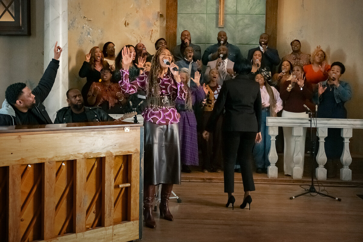 A gospel choir singing passionately in a church, with a pianist, soloist, conductor, and choir members standing before a cross, filling the space with music and energy.