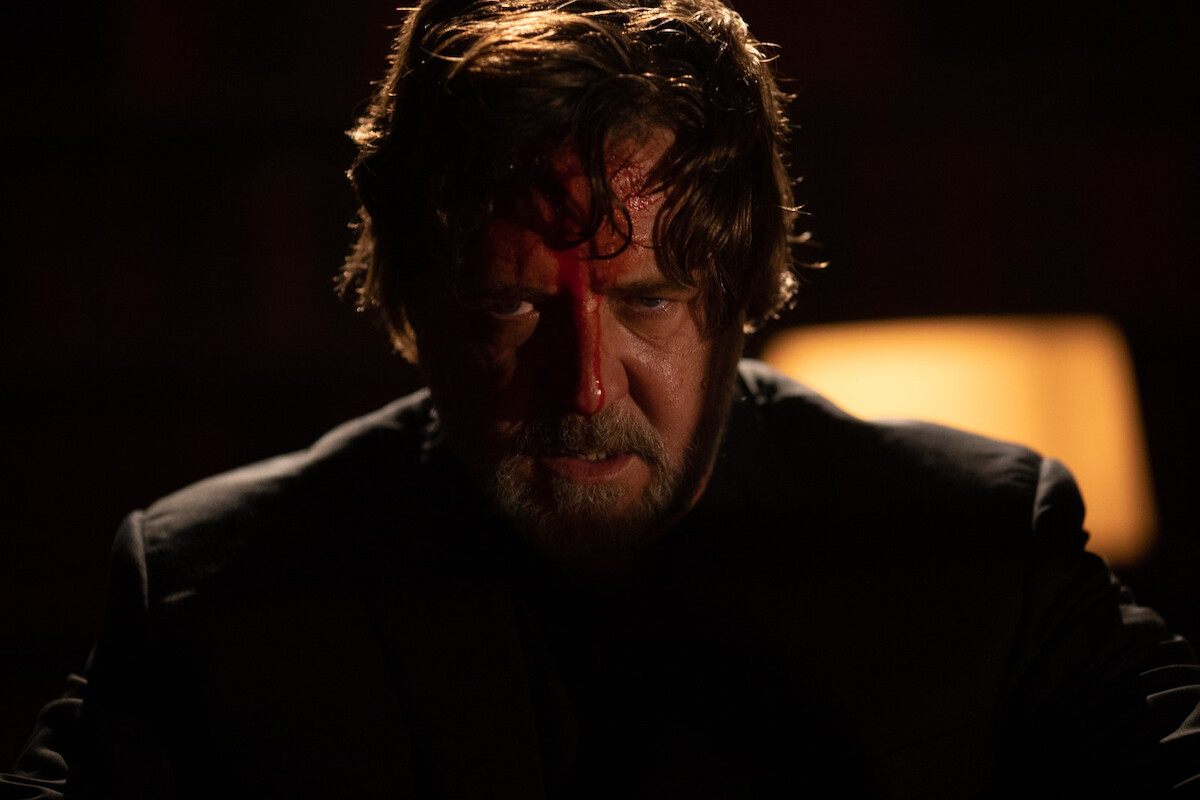 A man with disheveled hair and a serious expression, blood on his forehead, sits in a dimly lit room with a blurred lamp in the background, conveying a tense or dramatic atmosphere.
