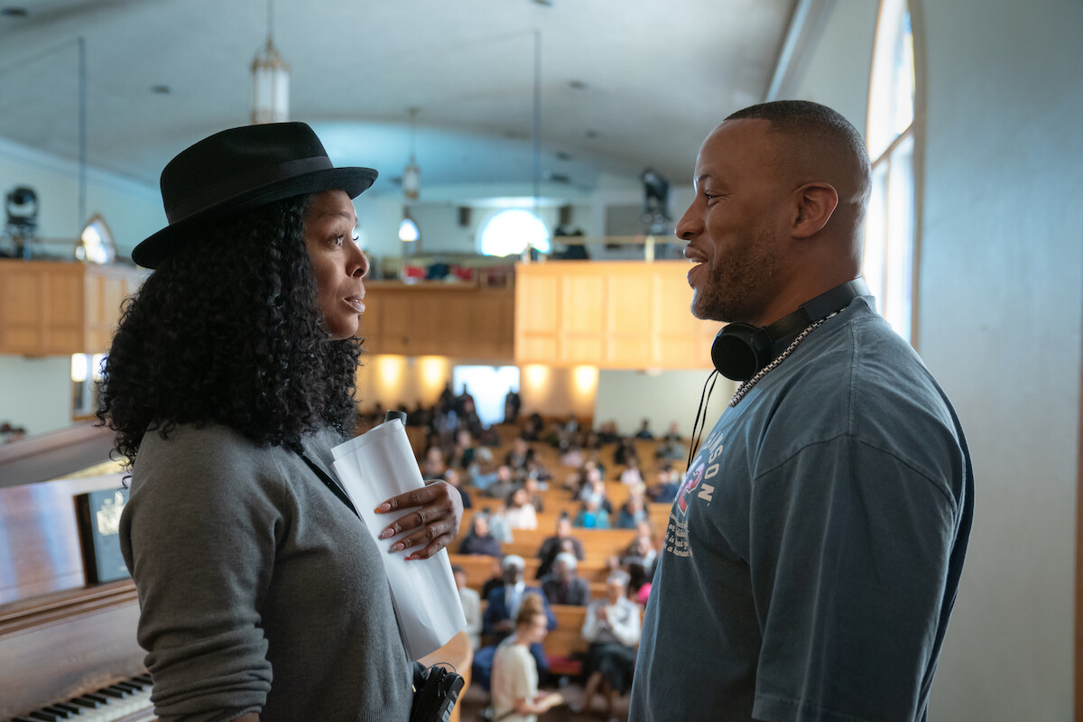 Two people have a conversation in a church or auditorium filled with people. One holds papers, the other wears headphones around his neck. Sunlight streams through windows, creating a warm, lively atmosphere.