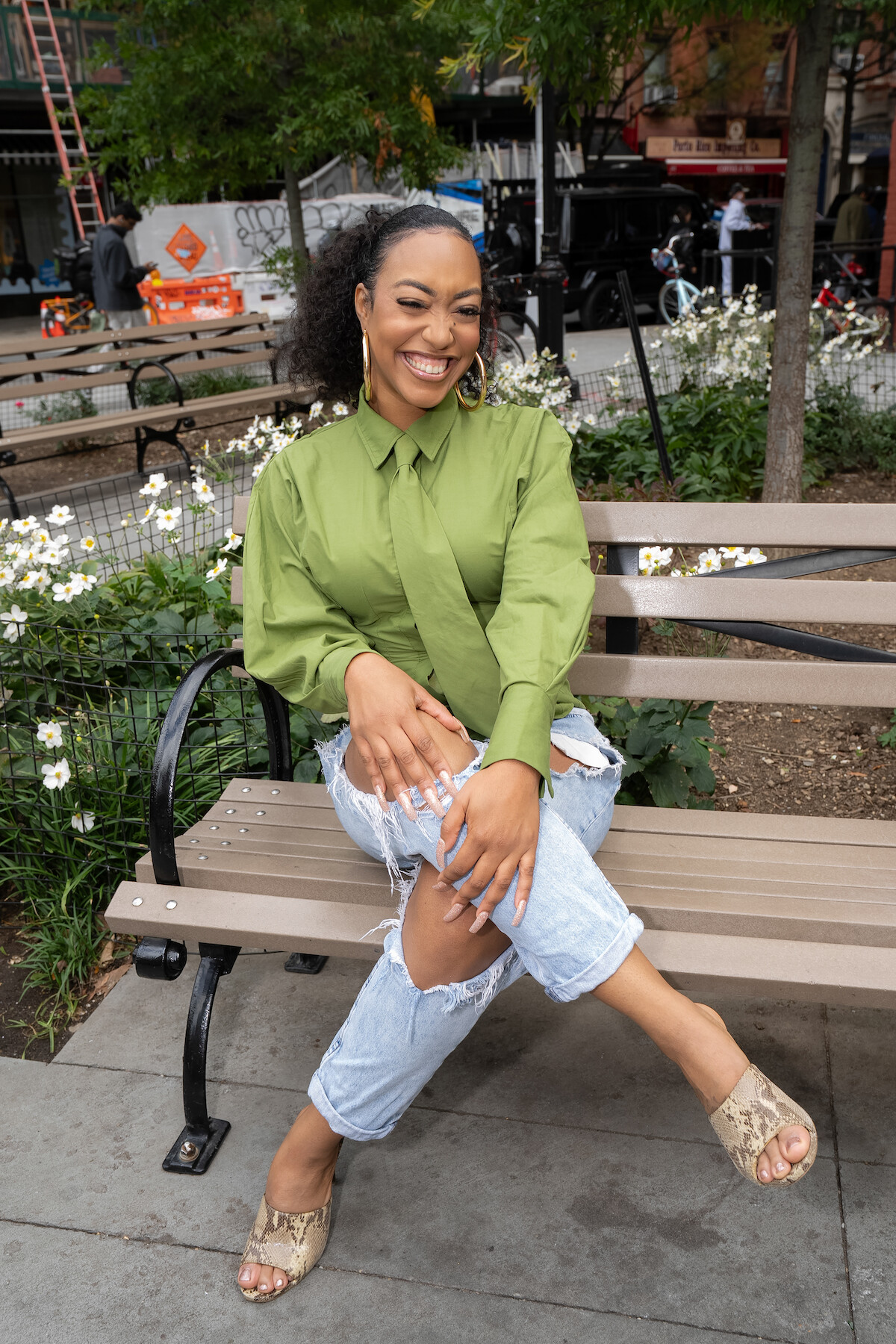 Smiling woman in green shirt and ripped jeans sitting on a park bench, garden flowers in background, city street scene with trees, people, and buildings visible around her.