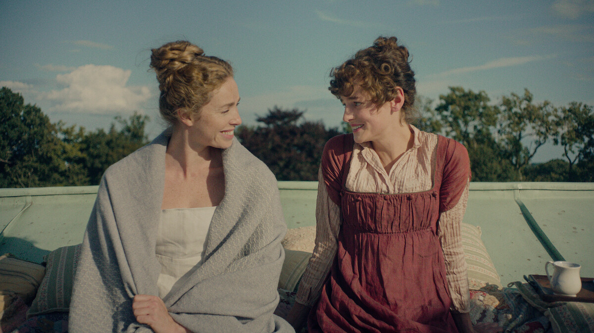 Two women in historical clothing sit outside on a rooftop, smiling at each other with greenery and blue sky in the background. Pillows and a cup are nearby, creating a relaxed, intimate setting.