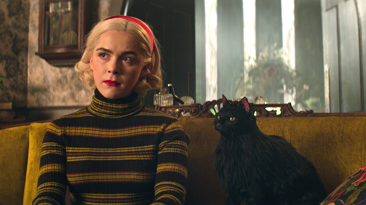 Young woman with short blonde hair and red headband sits on a vintage yellow sofa beside a black cat in a cozy, dimly lit room with patterned wallpaper and arched windows in the background.
