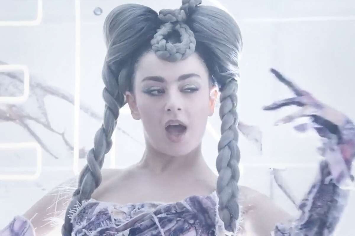 Woman with elaborate braided hairstyle and avant-garde makeup poses in a futuristic, softly lit environment, wearing a unique patterned outfit with feathery details. White neon light elements and abstract decor create a surreal, artistic setting.