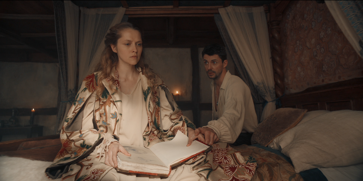A woman in ornate robes sits on a canopy bed reading a large book, while a man in historical clothing sits beside her. The room is warmly lit with candles, featuring wooden walls and patterned bedding, creating an old-fashioned atmosphere.