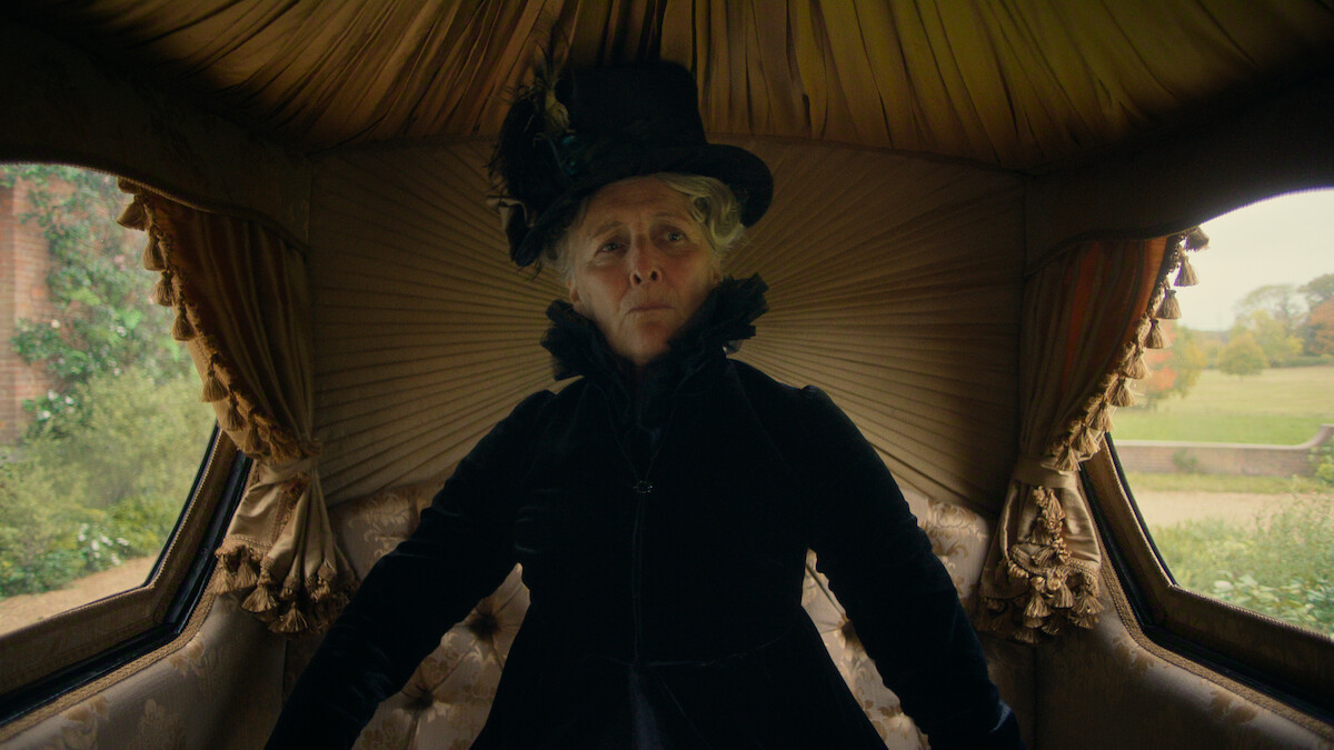 An older woman in Victorian-era black attire and hat sits sternly inside a plush, ornate carriage with draped gold fabric and window views of a garden or estate outside.