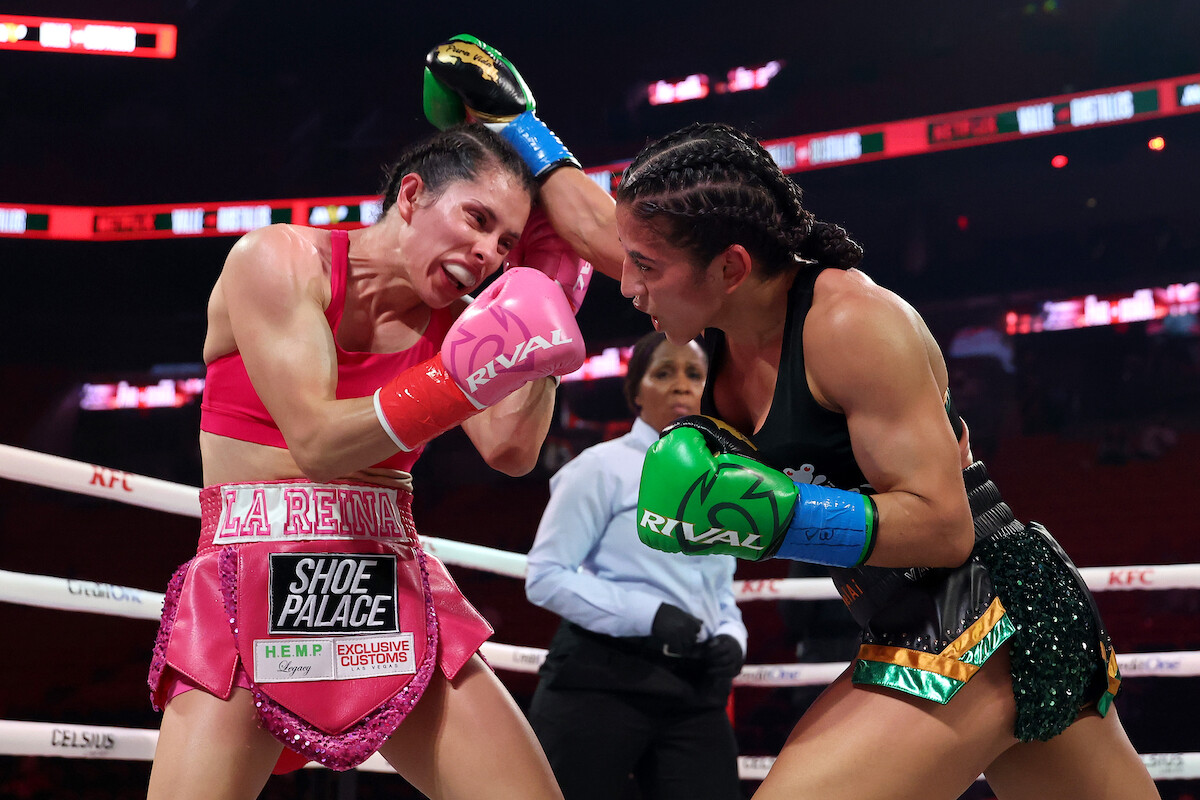 Yadira Bastillos and Yokasta Valle exchanging punches in a boxing ring during a match, wearing gloves and athletic gear, with a referee watching closely in the background and arena lights overhead.