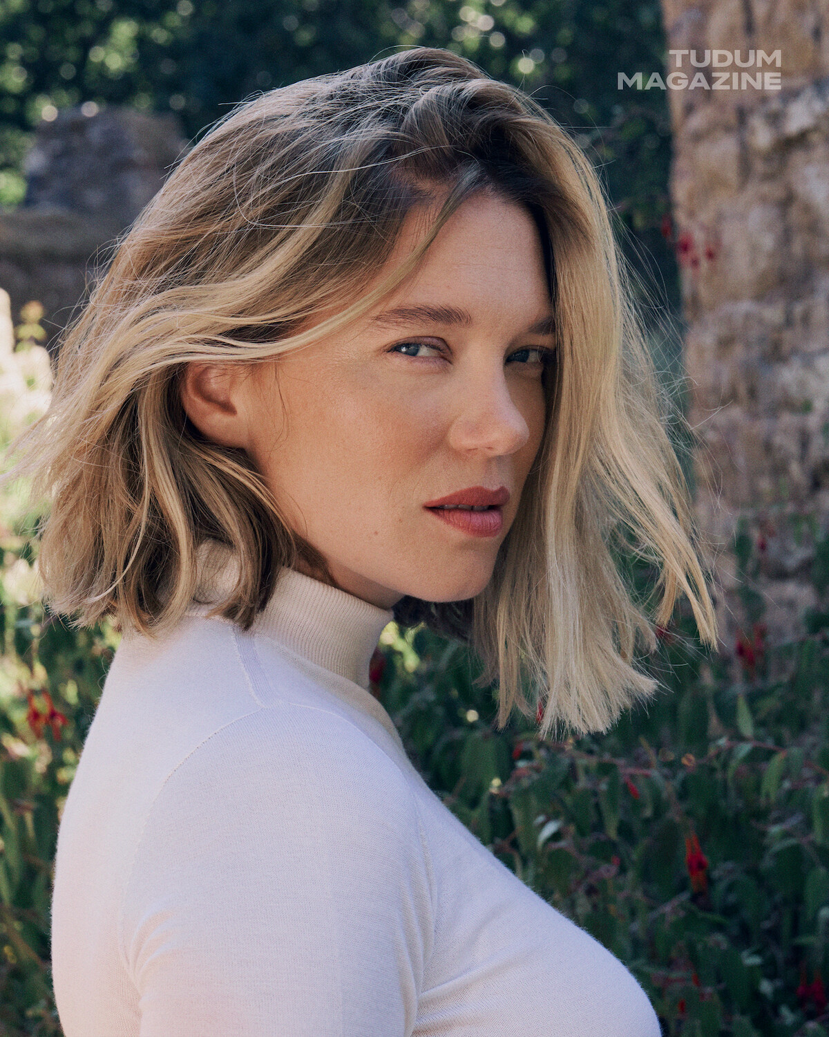 A woman with blonde hair wearing a white turtleneck stands outdoors in natural light, surrounded by greenery and stone, looking at the camera. The photo has a calm, contemplative mood.