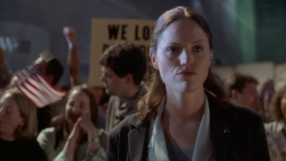 A woman stands in focus in the foreground at a crowded outdoor event, with people holding signs and small American flags in the background, indicating a public gathering or protest at night.