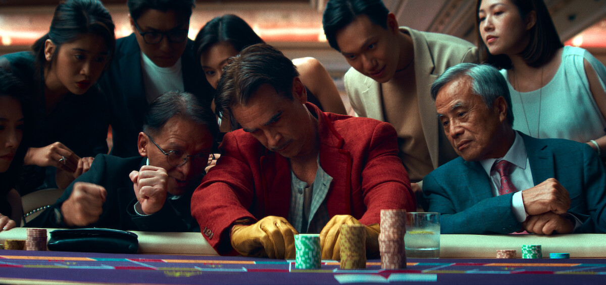A group of people gather intensely around a casino table covered with chips and drinks, creating a tense and suspenseful atmosphere. The main subject looks focused and serious, suggesting high stakes in a lively gambling environment.