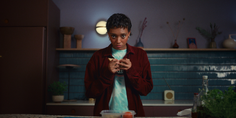 Sophie Wilde as Mia stands in a kitchen holding a piece of bread in Season 1 of 'Everything Now.'