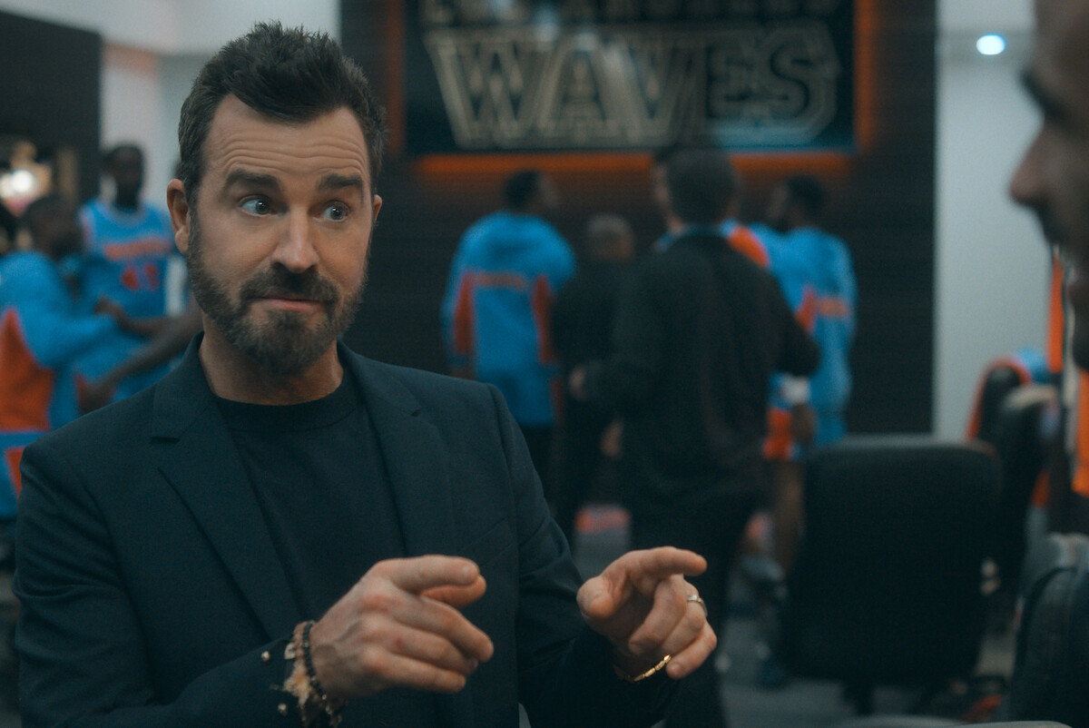 Man with beard gesturing animatedly indoors, sports team wearing blue and orange uniforms in the background, locker room with "WAVES" sign on the wall, casual and energetic atmosphere.