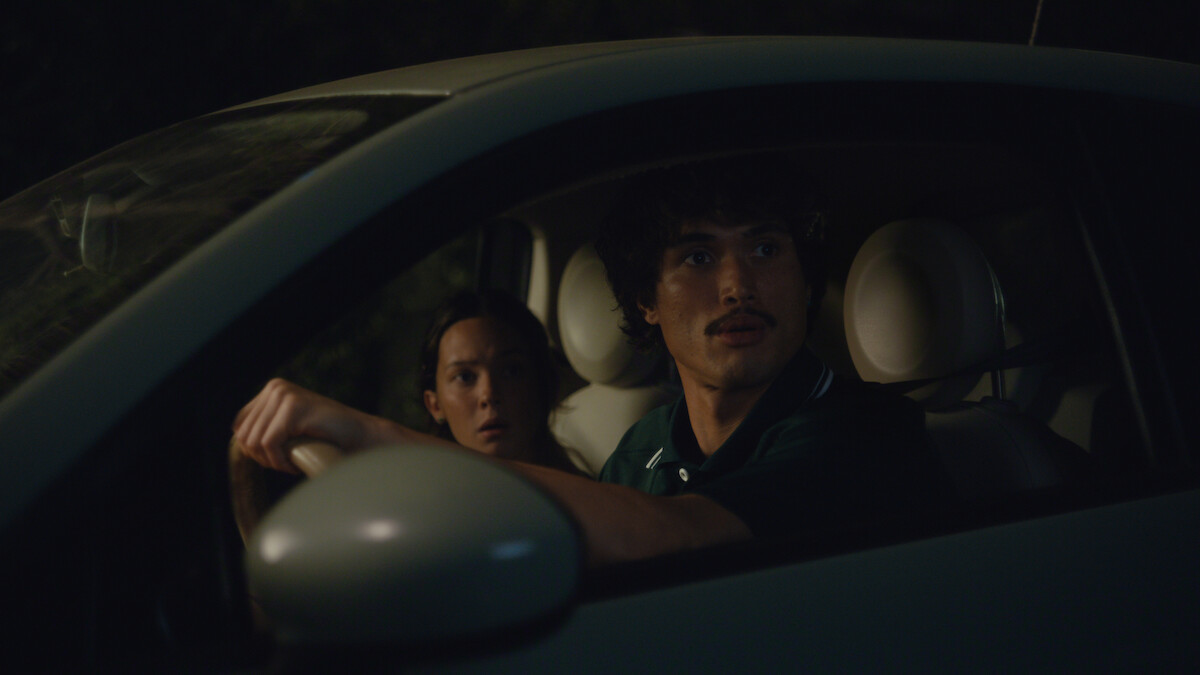 Two people sit in a small car at night, both looking out the window with surprised or concerned expressions, illuminated by dim light, with a dark outdoor background.