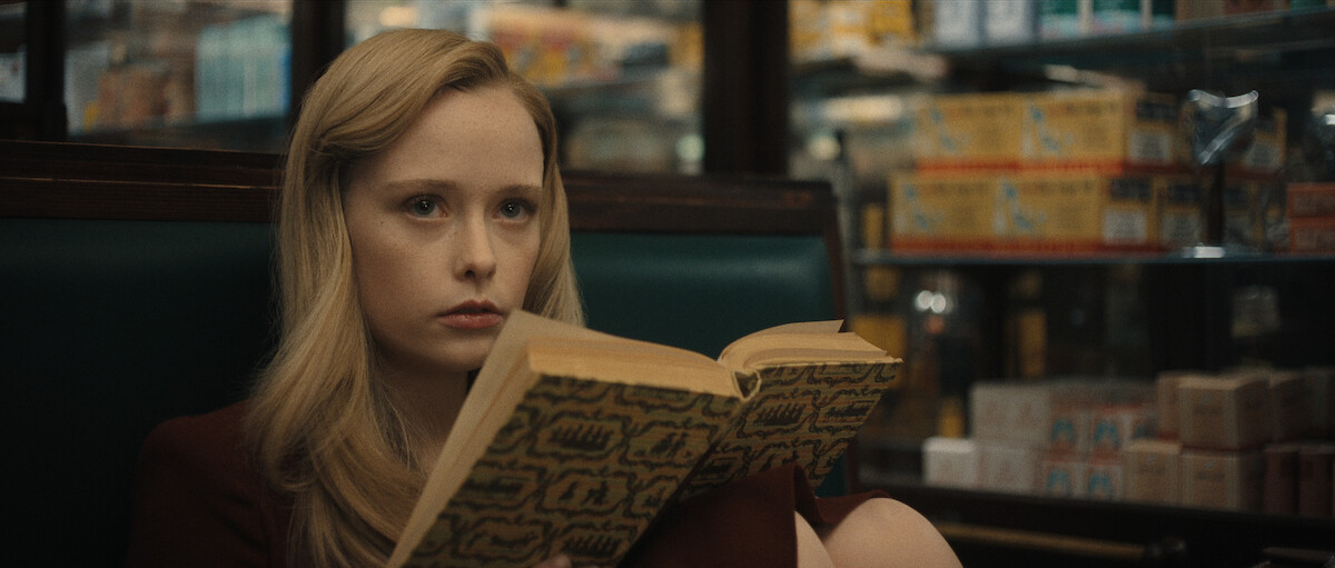 A young woman with long blonde hair sits in a booth, holding an open book and staring ahead with a serious expression in a store or diner setting.
