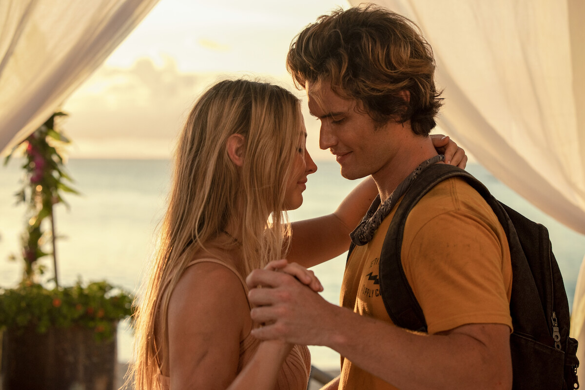 A young couple shares an intimate moment, holding hands and gazing at each other, standing on a beach at sunset under white draped fabric. The setting is romantic and warm with soft light and ocean in the background.