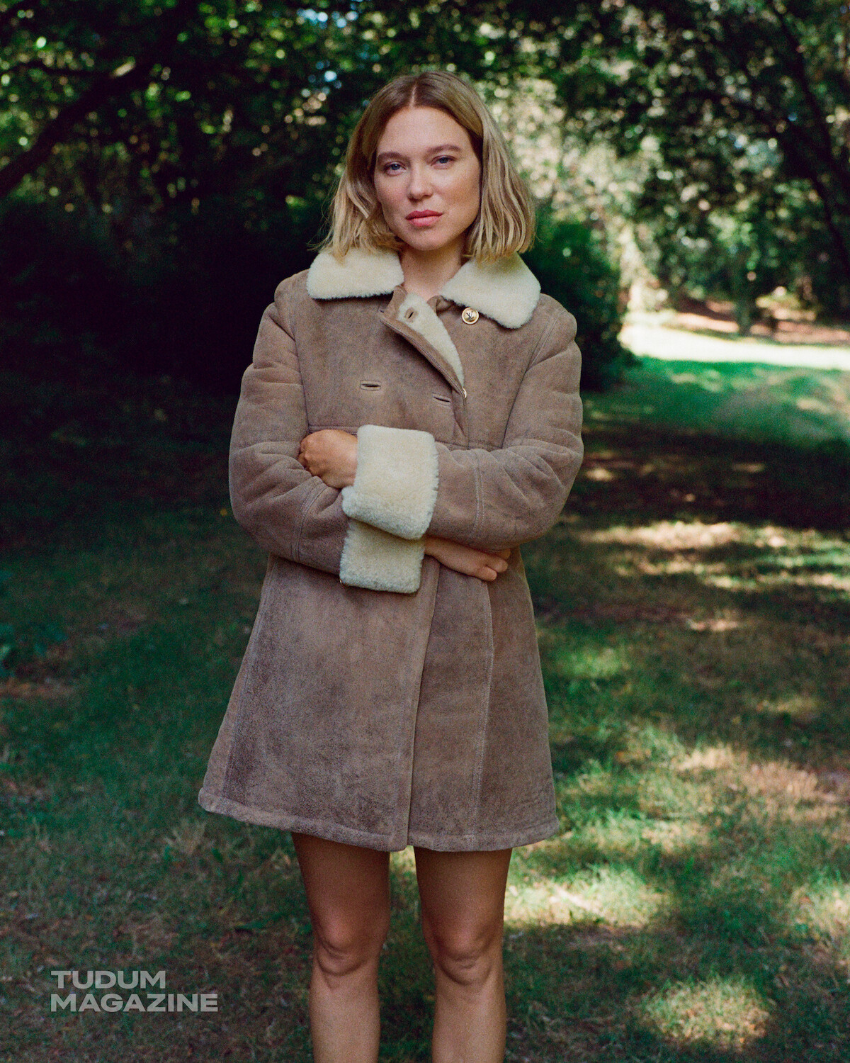 A woman in a brown shearling coat stands with arms crossed on a grassy area surrounded by trees in daylight. The setting is outdoors, with dappled sunlight filtering through the foliage. Tudum Magazine logo appears in the corner.