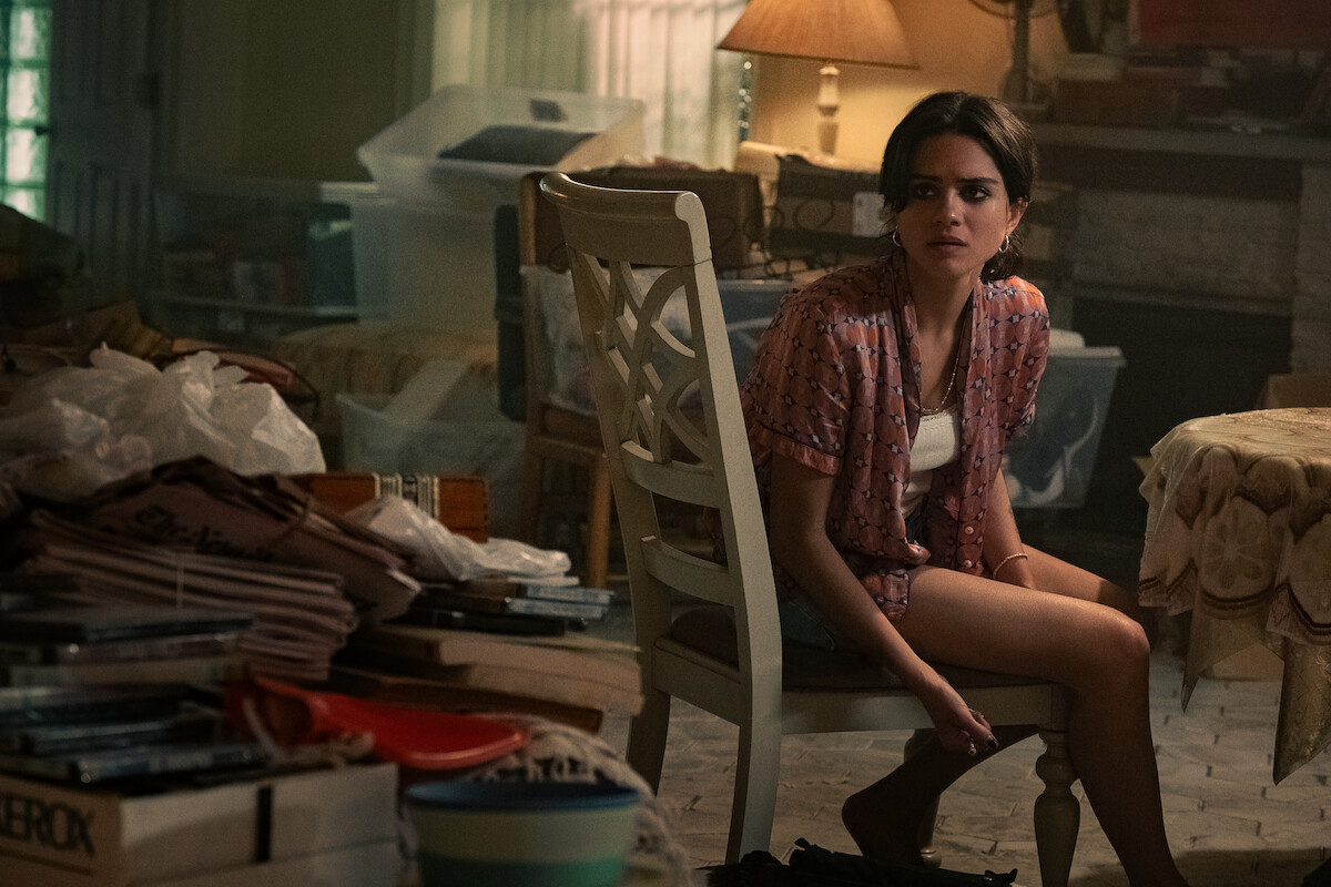 Young woman sitting on a chair in a cluttered, dimly lit room filled with stacks of papers, boxes, and covered furniture, with warm lighting from a table lamp and a window in the background.