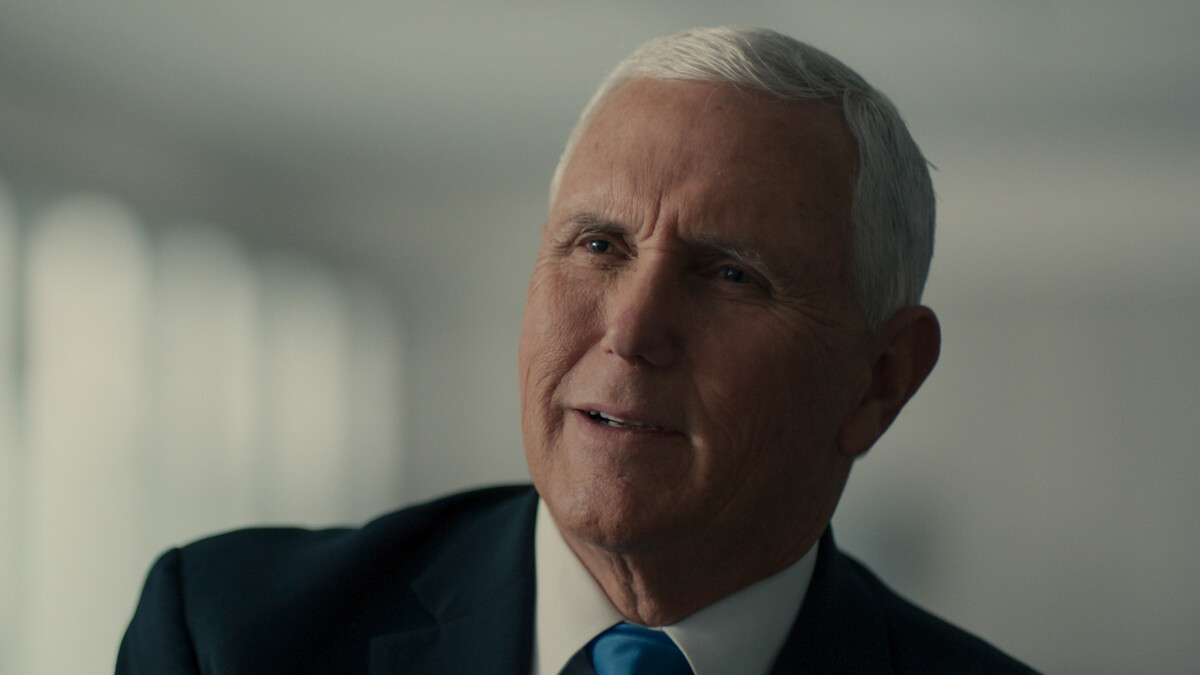 Older man with short gray hair wearing a suit and blue tie, seated indoors in a softly lit room with blurred windows in the background, looking slightly off camera with a thoughtful expression.