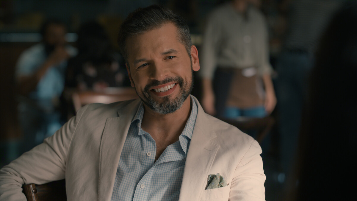 A man with a beard, wearing a light blazer and checked shirt, smiles while sitting at a table in a softly lit café, with blurred people and a casual atmosphere in the background.