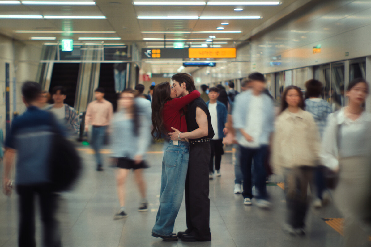 A couple embracing and kissing in the middle of a busy subway station, surrounded by blurred commuters walking past them under bright fluorescent lights.