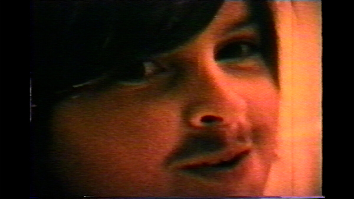 A close-up, grainy image of Charlie Sheen as a child with long dark hair, a fake mustache, and a slight smile, in warm lighting.