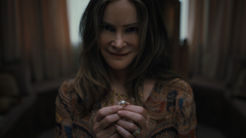 A woman with long brown hair and patterned blouse smiles subtly while holding a ring in both hands, in a dimly lit room with soft curtains and neutral furniture in the background.