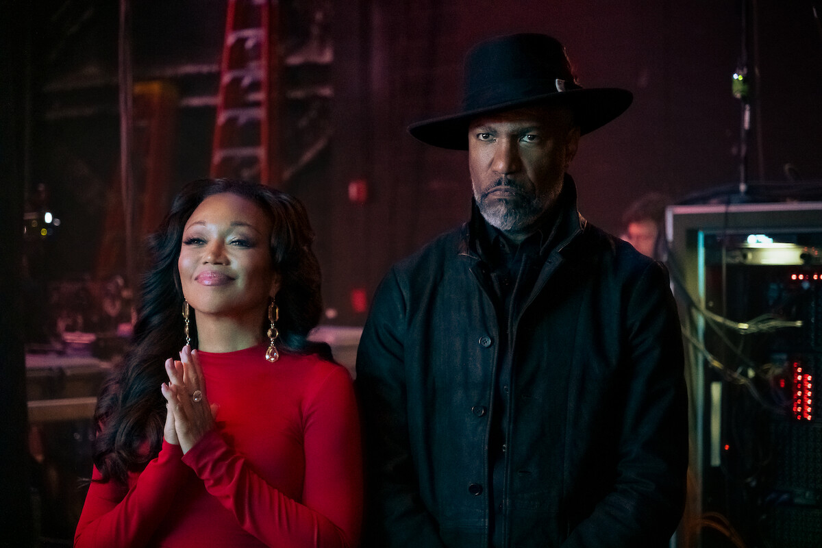 A smiling woman in a red dress and a serious man in a black coat and hat stand side by side backstage, with ladders and technical equipment visible in the dimly lit background.