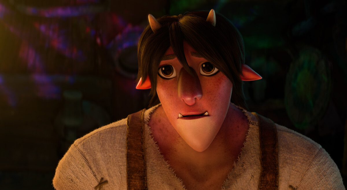 Animated character with small horns, pointy ears, and a concerned expression, wearing a rough shirt and suspenders, in a dimly lit, rustic environment with colorful light reflections on the wall behind.
