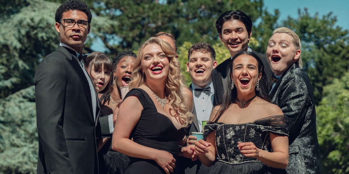 Group of young adults in formal attire posing outdoors, smiling and laughing together for a photo, with green trees and blue sky in the background, suggesting a celebratory event like prom or a formal party.