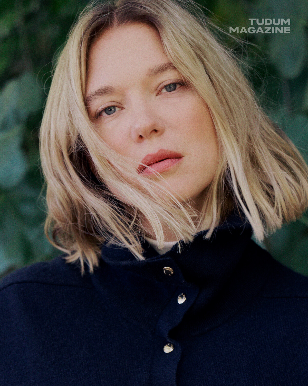 Woman with blonde hair in a dark buttoned sweater, posing outdoors in front of green foliage, with soft natural light. "Tudum Magazine" text in the upper right corner.
