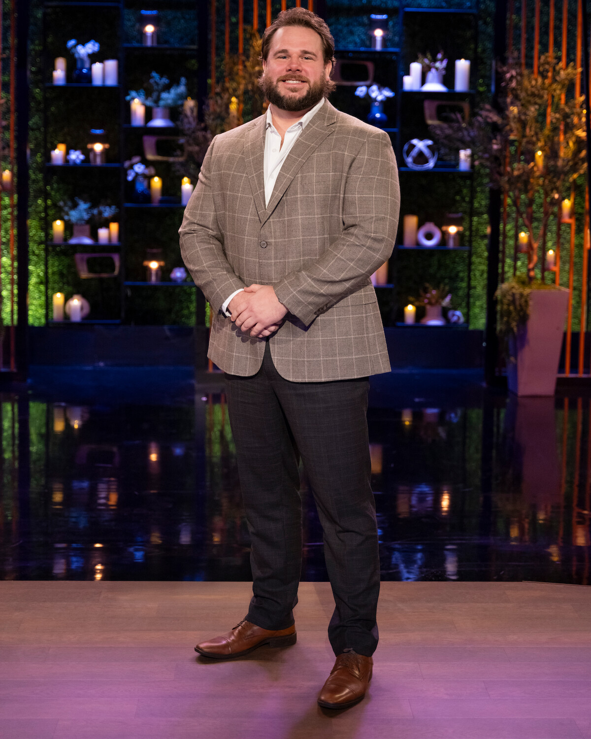 A man in a checked suit jacket stands smiling on a stage with decorative shelves behind him, featuring plants and candles, in a warmly lit indoor setting.