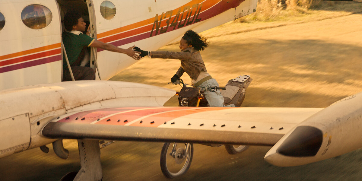 Uma mulher em uma moto estende a mão para agarrar a mão de um homem pela porta aberta de um avião, enquanto a aeronave decola em uma pista de terra, criando uma cena de ação dramática ao ar livre.