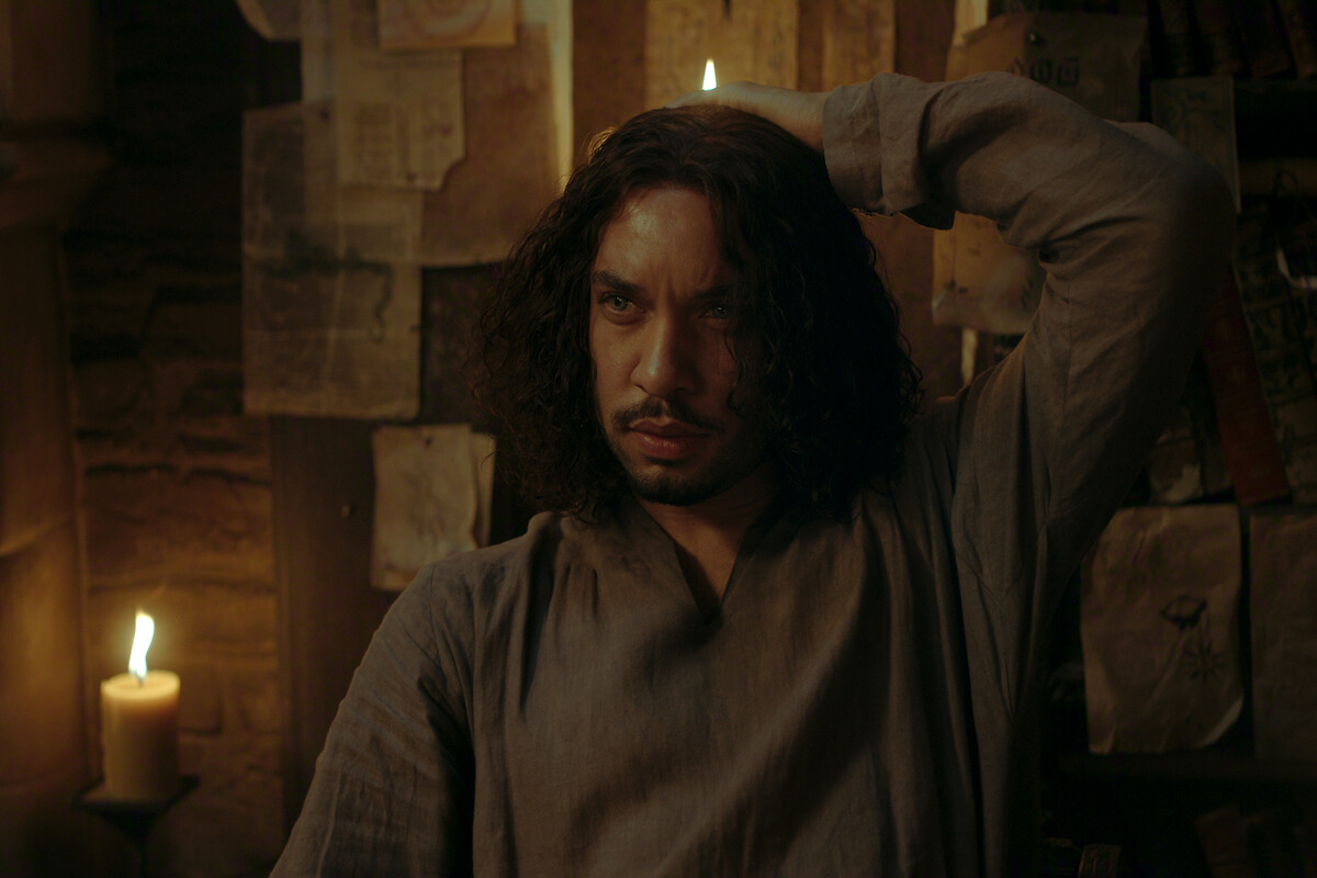 A man with long hair sits in a dimly lit room, surrounded by candles and old papers on stone walls, creating a moody and contemplative atmosphere.