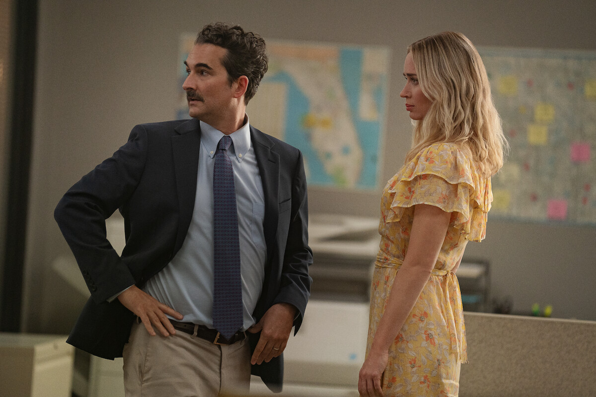 Jay Duplass stands with his hands on his hips next to an upright Emily Blunt in ‘Pain Hustlers.’