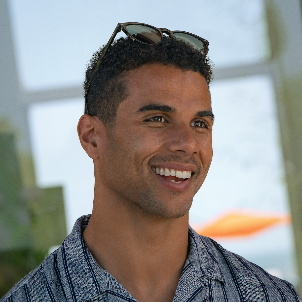 Mason Gooding as Mason with short curly hair and sunglasses on his head, wearing a striped shirt. He is indoors with sunlight and blurred outdoor scenery, possibly a beach or waterfront, visible through windows behind him. Relaxed, casual setting.