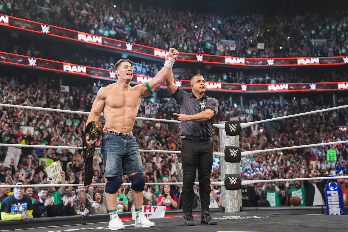 A muscular man in a red shirt and cap stands confidently in a wrestling ring holding a microphone, with a lively crowd in the background, creating an energetic and celebratory atmosphere at a professional wrestling event.