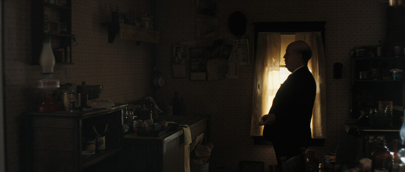 A man in a suit stands in a dimly lit, cluttered kitchen, facing a window with light streaming through the curtains.