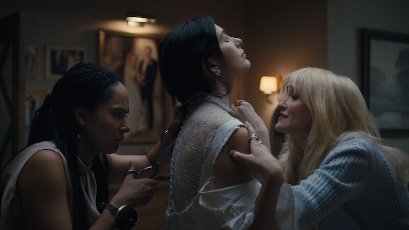 Three women in a dimly lit room, one holding scissors cut the back of another’s white lace dress while a third woman appears to comfort or steady her, with framed art and soft lamp lighting in the background.