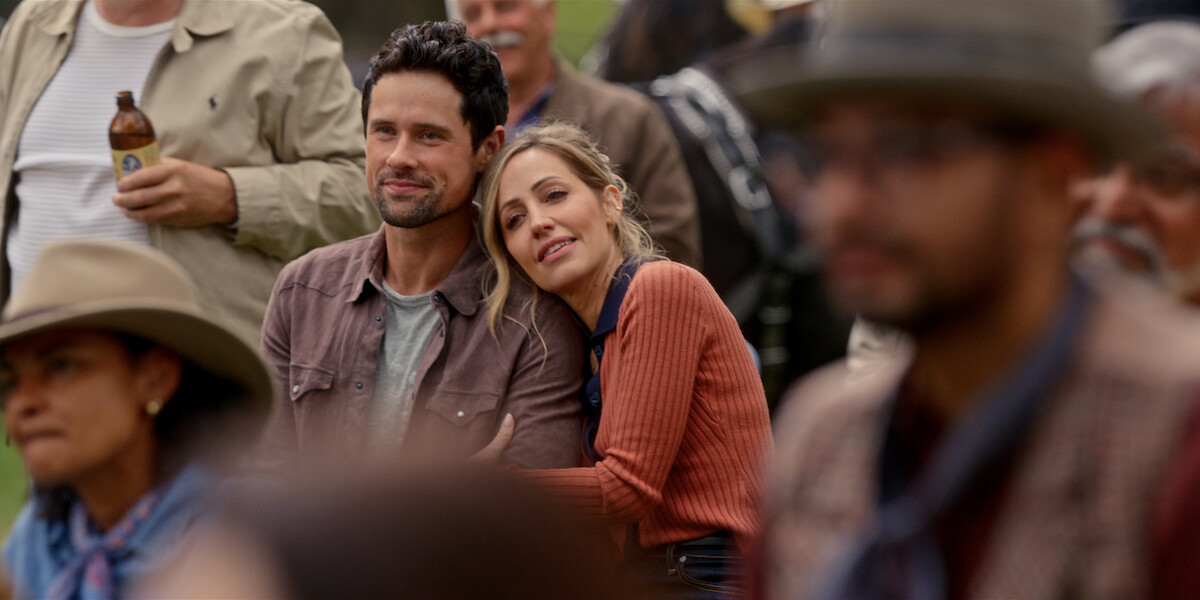 A couple sits closely together at an outdoor gathering, surrounded by other people in casual and western-style clothing, creating a warm, relaxed atmosphere.