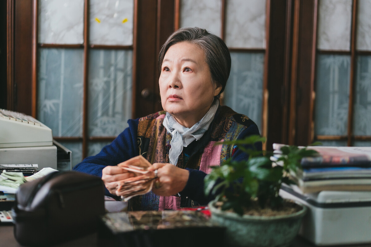 Elderly woman sits at a desk counting money, surrounded by office supplies, a calculator, papers, and a potted plant, in a traditional room with wooden and frosted glass panels.