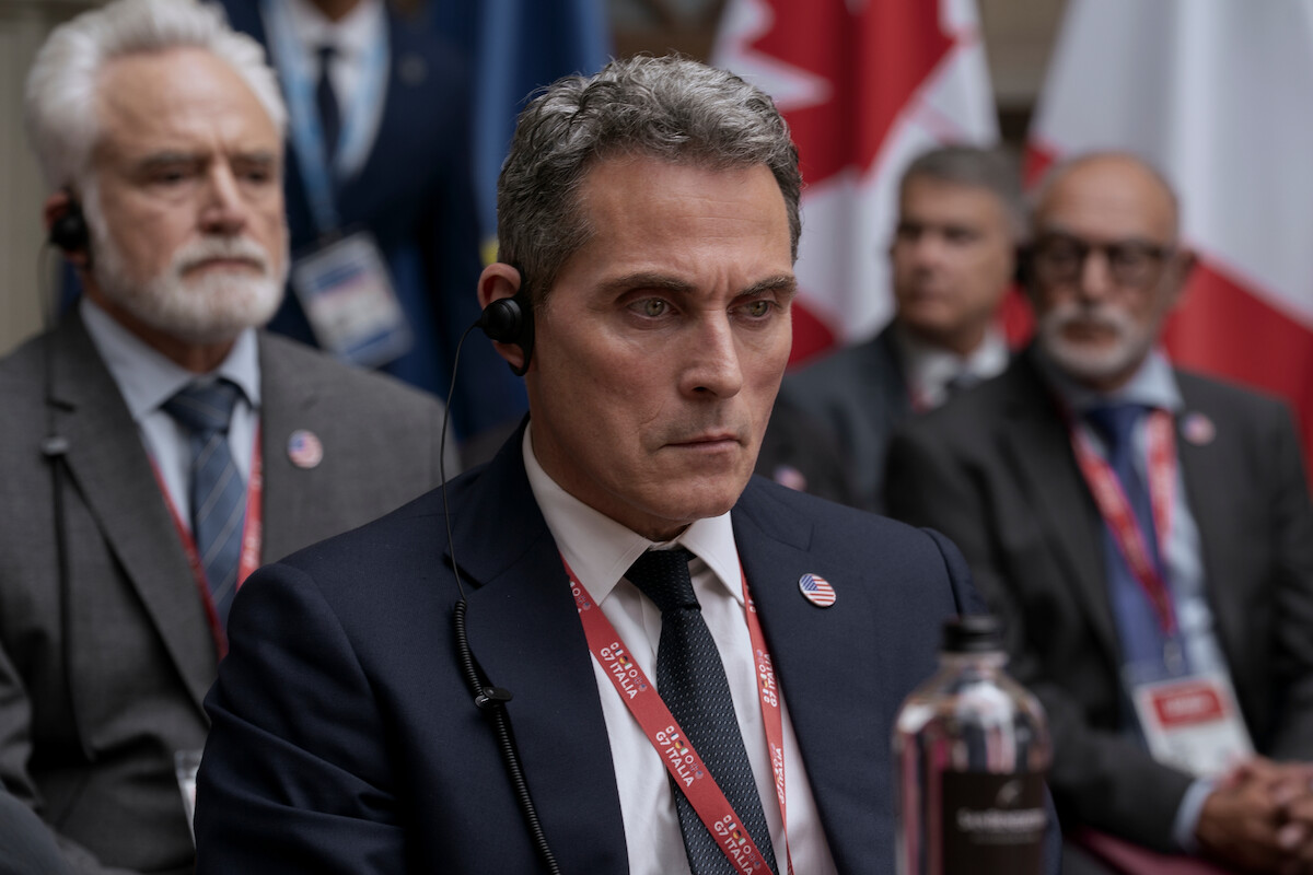 Serious men in suits with headsets sit at a formal international conference, wearing badges and lanyards, with national flags in the background, suggesting a diplomatic or government meeting setting.