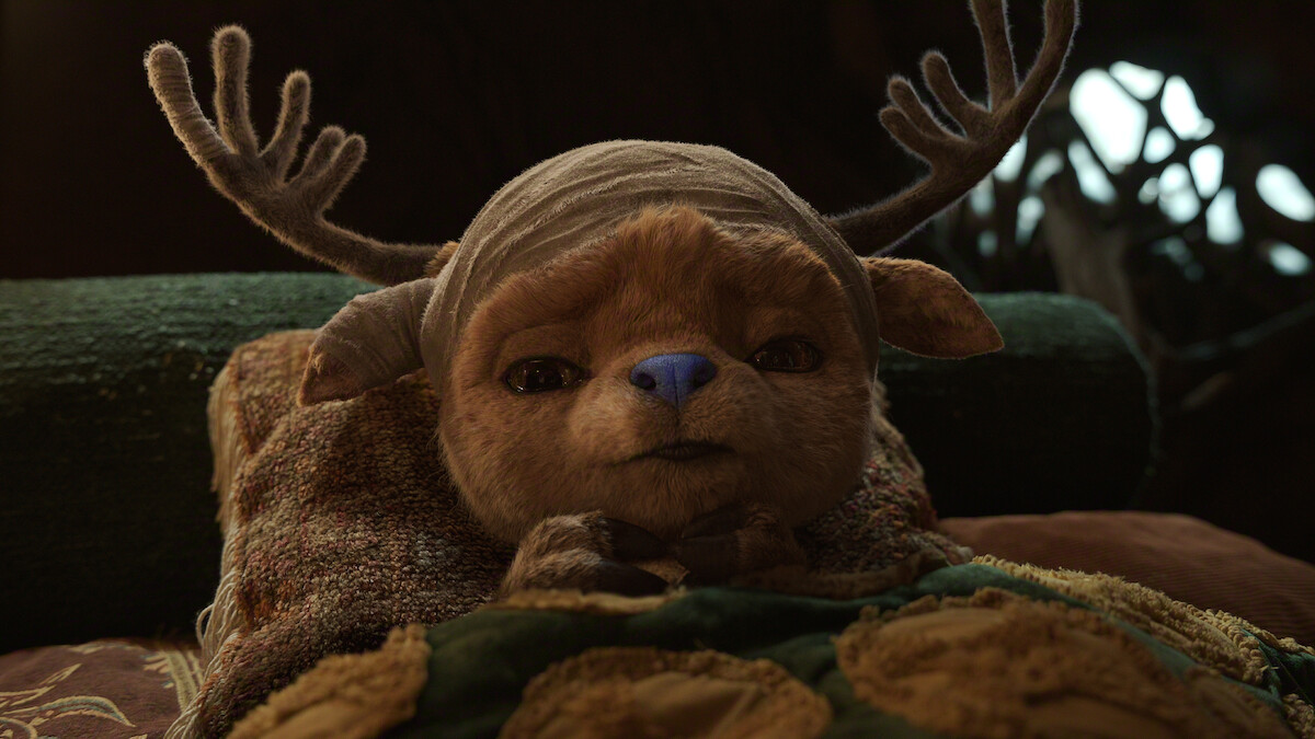 Tony Tony Chopper, a small creature with antlers and a bandaged head lies in bed surrounded by pillows and blankets in a cozy, dimly lit environment.