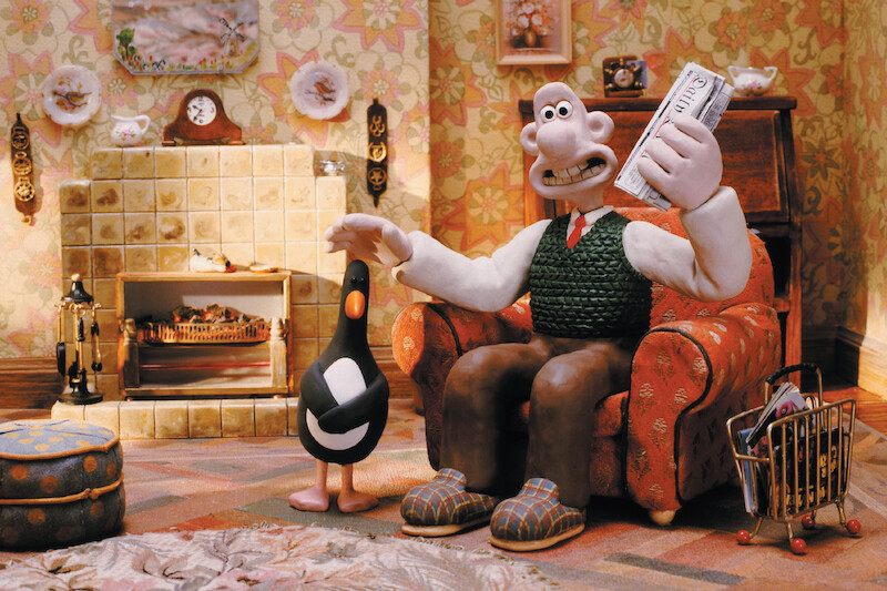Wallace wears brown pants, a white shirt, and green vest in his living room.