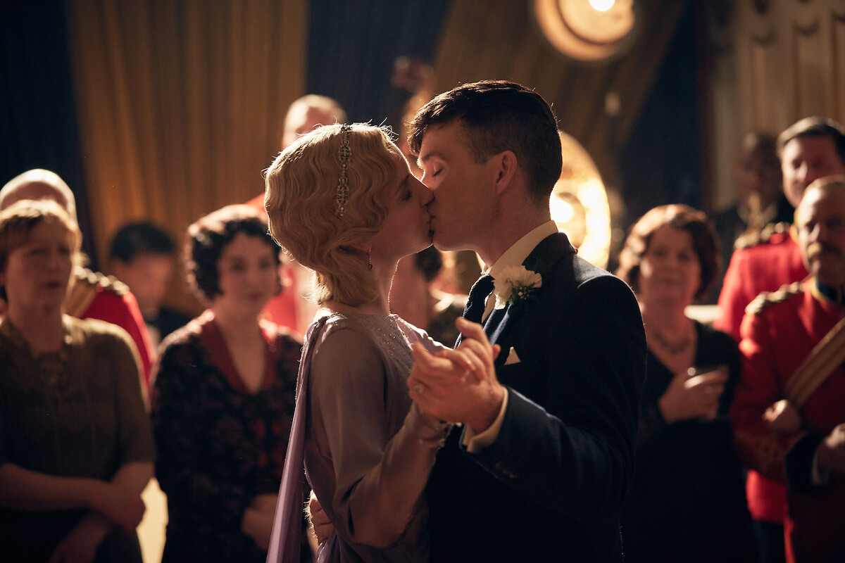 A couple in formal attire shares a kiss while dancing at a vintage-themed party or wedding, surrounded by elegantly dressed guests in a warmly lit, classic ballroom setting.