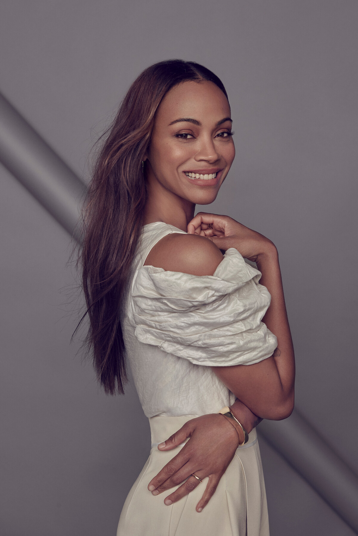 Zoe Saldaña, facing the camera, smiles in her cream outfit that has a dramatic textured shoulder element.