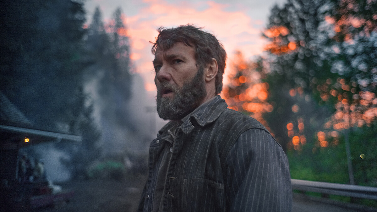 Bearded man in rugged clothing stands outdoors at dusk, with a dramatic sunset, trees, and smoke in the background, creating a tense and contemplative mood.