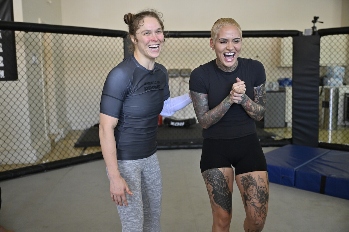 Ronda Rousey and Kehlani laughing together in an MMA training gym, standing on mats near a fenced cage, wearing athletic workout clothing with one sporting visible tattoos.