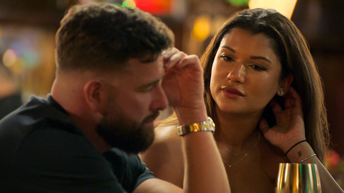 Two people sitting close together at a dimly lit bar or restaurant, engaged in a serious conversation. The man looks contemplative, while the woman listens attentively, touching her hair. Warm, intimate setting with soft background lights.