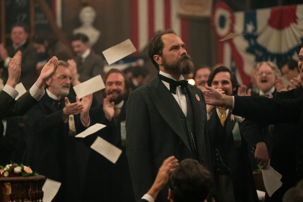 A bearded man in 19th-century formal attire stands serious in a crowded hall, surrounded by clapping, cheering men and flying papers, with American flags and patriotic decorations in the background.