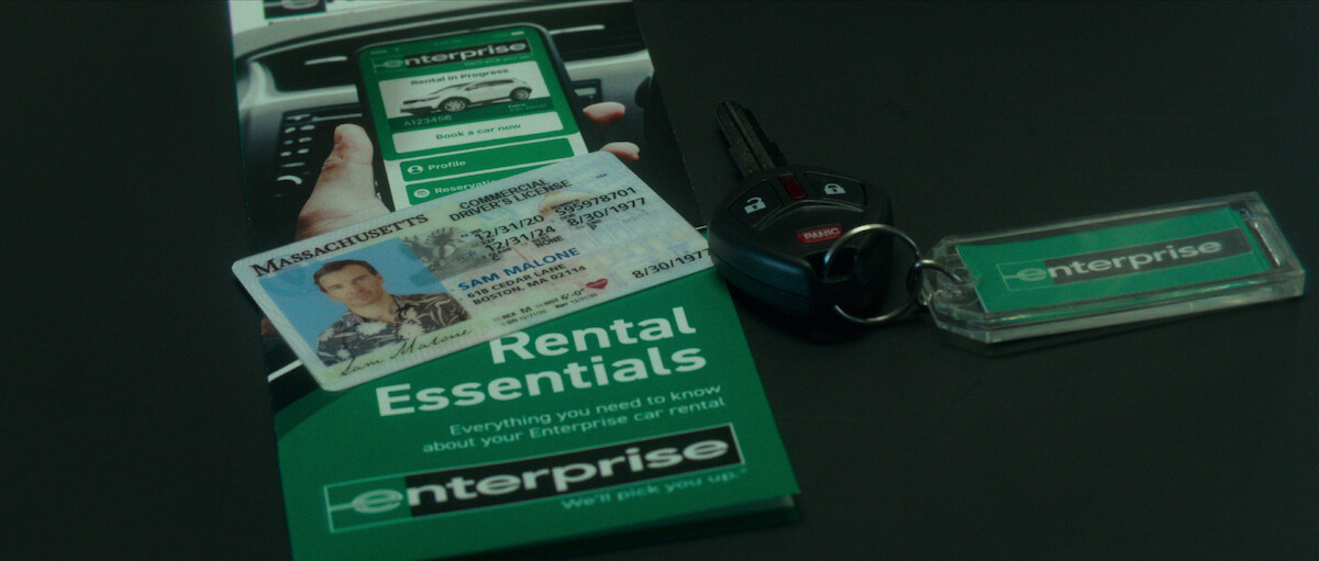 A Massachusetts driver’s license that reads Sam Malone sits on top of an Enterprise car rental pamphlet and beside a car key.